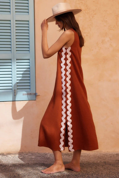 Brown - Full-length view of a woman in a wide-brimmed hat and a sleeveless, rust-orange midi shift dress featuring a white wavy trim down the side. She stands barefoot against a peach wall with blue shutters.