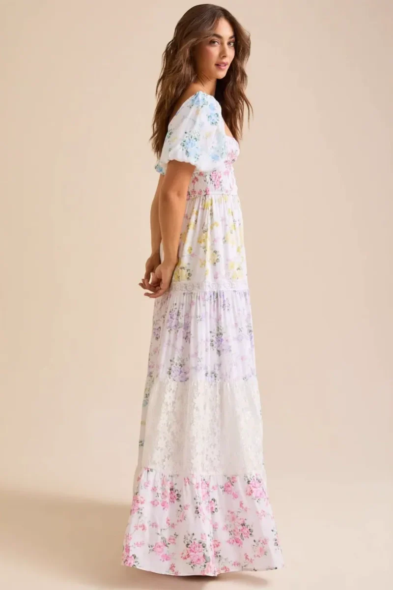 Colorful - A woman wearing a white, tiered maxi dress with a smocked bodice, short puff sleeves, and a pastel patchwork floral print with lace inserts.