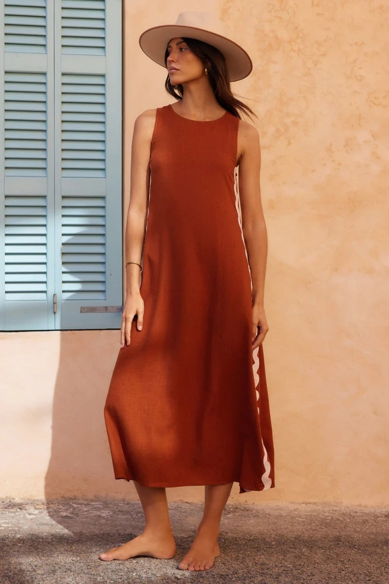 Brown - Full-length view of a woman in a wide-brimmed hat and a sleeveless, rust-orange midi shift dress featuring a white wavy trim down the side. She stands barefoot against a peach wall with blue shutters.