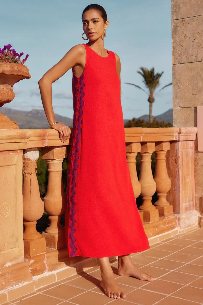 Brown - Full-length view of a woman in a wide-brimmed hat and a sleeveless, rust-orange midi shift dress featuring a white wavy trim down the side. She stands barefoot against a peach wall with blue shutters.