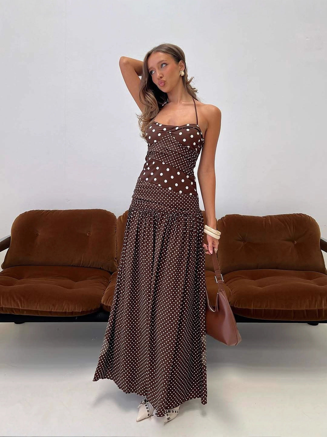Cocoa - A woman wearing a brown maxi dress featuring a mixed white polka dot print, a halter neckline, a fitted panelled bodice, and a flowing gathered skirt.
