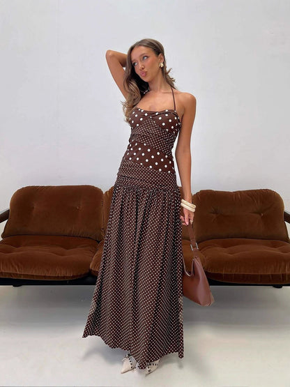 Cocoa - A woman wearing a brown maxi dress featuring a mixed white polka dot print, a halter neckline, a fitted panelled bodice, and a flowing gathered skirt.