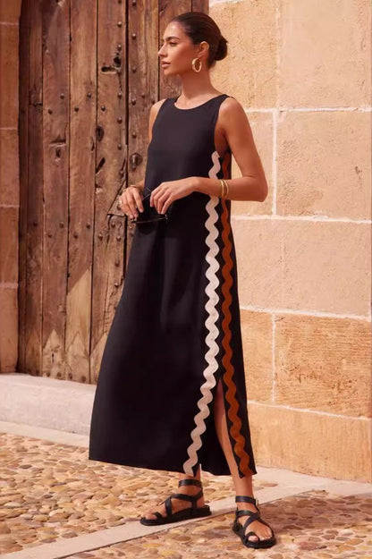 Black - Full-length view of a woman in a wide-brimmed hat and a sleeveless, rust-orange midi shift dress featuring a white wavy trim down the side. She stands barefoot against a peach wall with blue shutters.