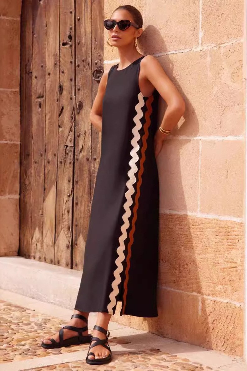 Black - Full-length view of a woman in a wide-brimmed hat and a sleeveless, rust-orange midi shift dress featuring a white wavy trim down the side. She stands barefoot against a peach wall with blue shutters.