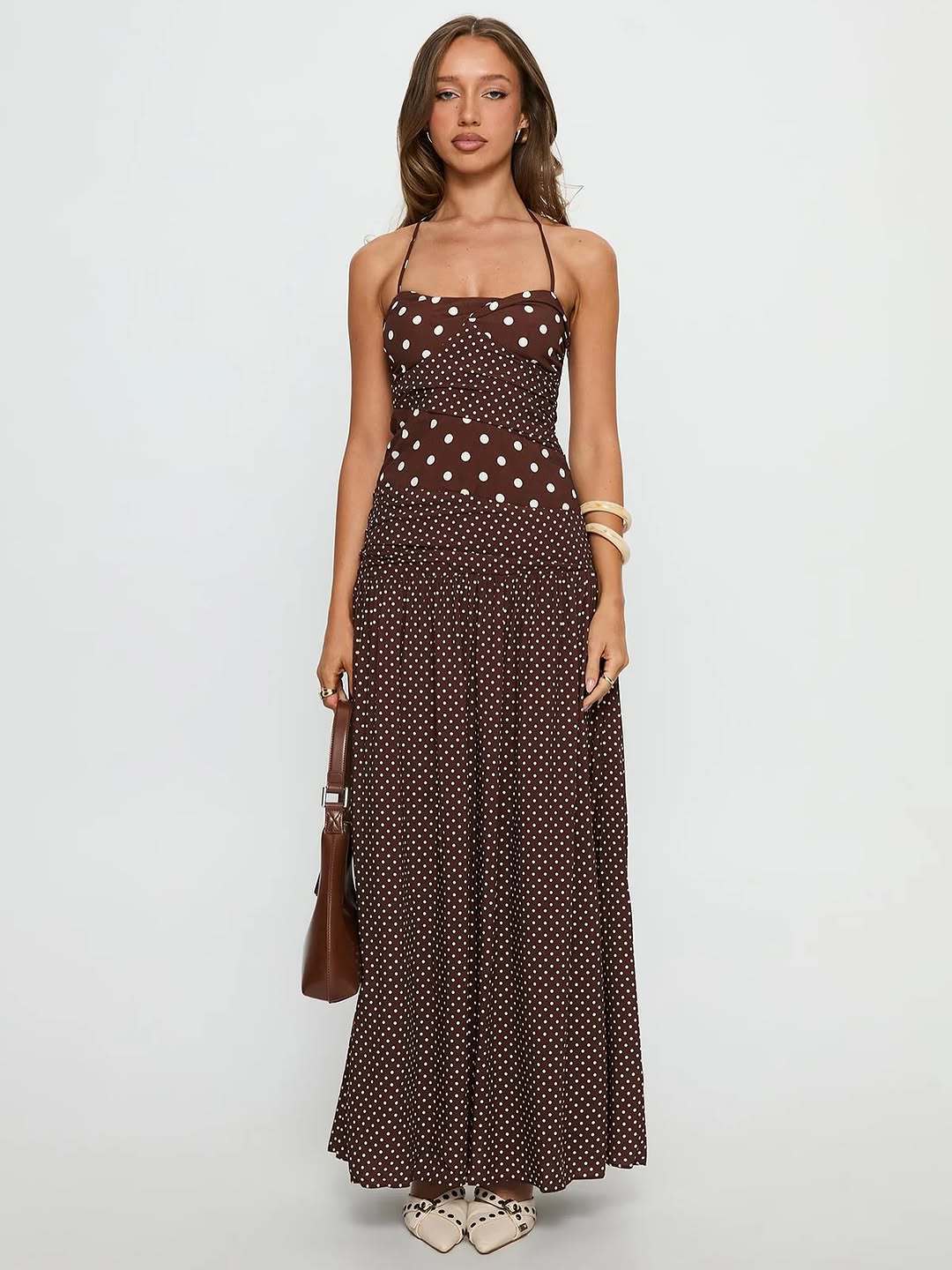 Cocoa - A woman wearing a brown maxi dress featuring a mixed white polka dot print, a halter neckline, a fitted panelled bodice, and a flowing gathered skirt.