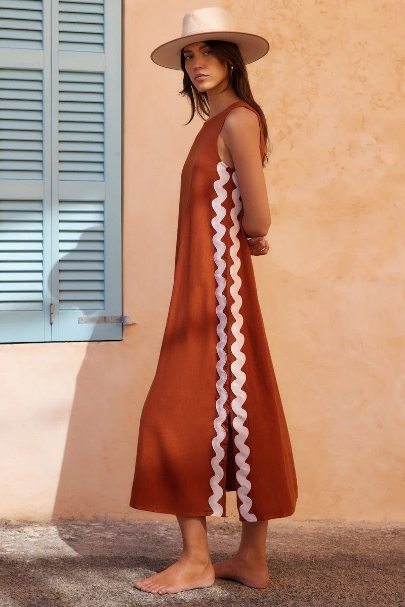 Brown - Full-length view of a woman in a wide-brimmed hat and a sleeveless, rust-orange midi shift dress featuring a white wavy trim down the side. She stands barefoot against a peach wall with blue shutters.