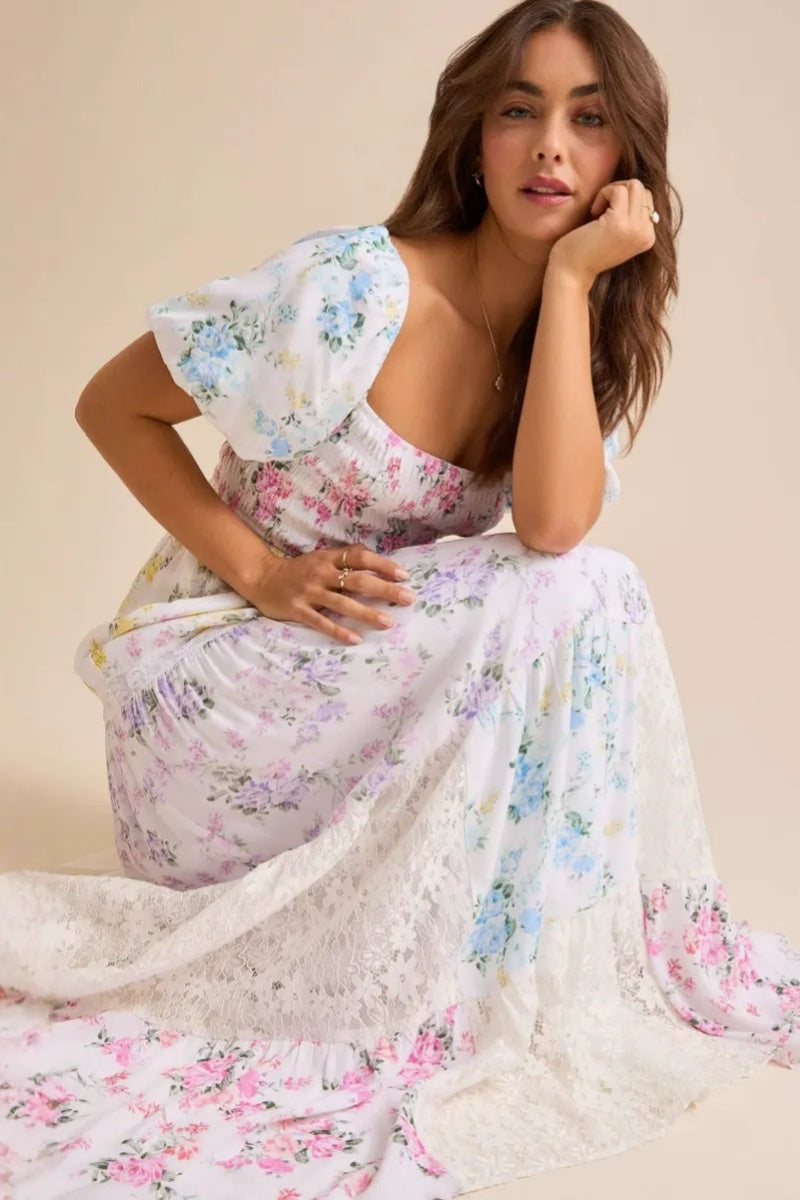 Colorful - A woman wearing a white, tiered maxi dress with a smocked bodice, short puff sleeves, and a pastel patchwork floral print with lace inserts.
