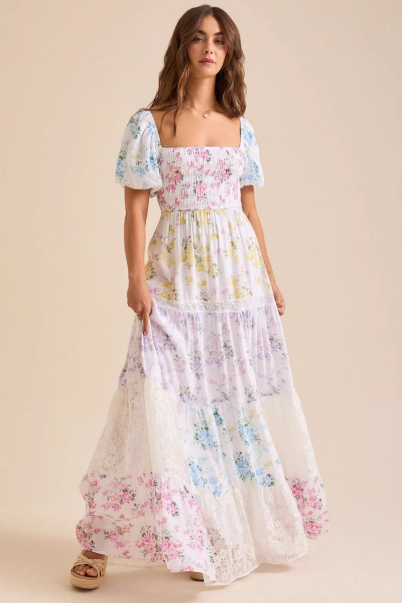 A woman wearing a white, tiered maxi dress with a smocked bodice, short puff sleeves, and a pastel patchwork floral print with lace inserts.
