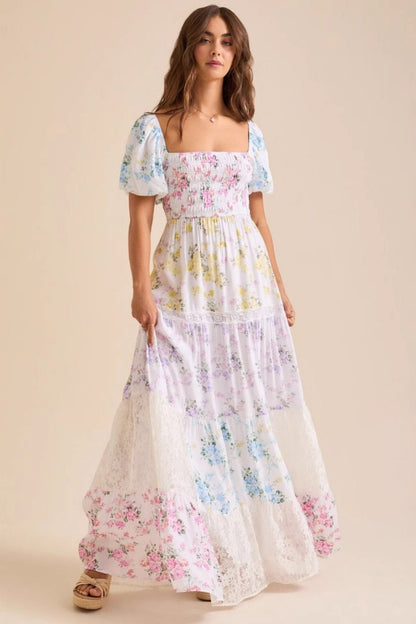 A woman wearing a white, tiered maxi dress with a smocked bodice, short puff sleeves, and a pastel patchwork floral print with lace inserts.