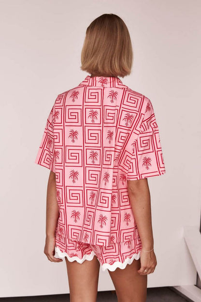 Pink - Model wearing a pink and red geometric palm print matching co-ord set featuring a short-sleeve button-up shirt and shorts with white scalloped trim.