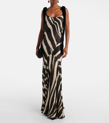 A woman modelling a floor-length, satin-look maxi dress featuring a bold, blurred zebra print and contrasting black textured shoulder ties.