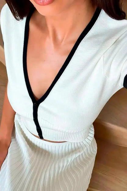 White - Woman wearing a white ribbed knit two-piece set featuring a short-sleeve V-neck crop top and a pleated midi skirt, both finished with black contrast trim.