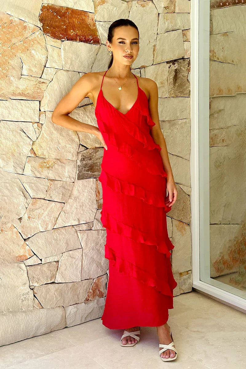 Red - A woman modelling a vibrant red, floor-length maxi dress featuring a V-neckline, thin straps, and elegant asymmetrical cascading ruffles down the front, standing against a light textured stone wall.
