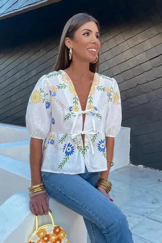 Women's white short-sleeve blouse featuring vibrant blue and yellow floral embroidery, double tie-front bow closures, and voluminous puff sleeves.