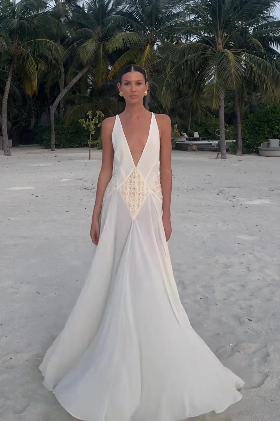 Ivory - A woman wearing an elegant, flowing off-white maxi dress featuring a deep plunging V-neckline and a textured appliqué waist panel, standing against a neutral background.