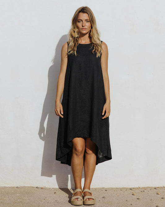 Black - A woman wearing a relaxed, black sleeveless trapeze midi dress with a high-low hemline and round neckline, standing against a white textured wall.