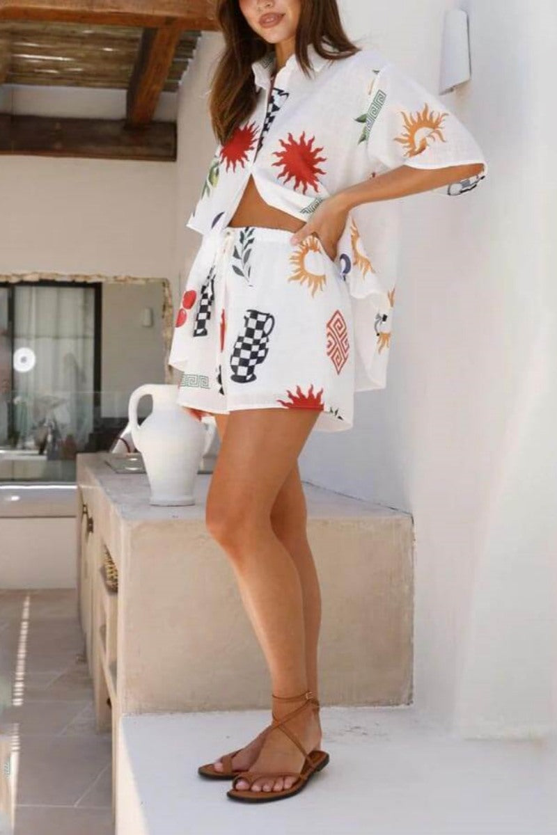 White - Women's white short-sleeved button-down shirt featuring a colourful Mediterranean motif print including suns, checkered vases, and evil eyes, worn partially unbuttoned over matching drawstring shorts.