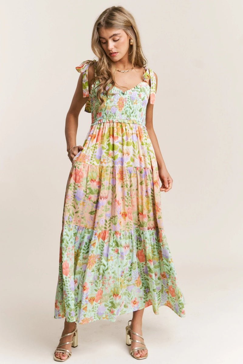 A woman wearing a vibrant floral print maxi dress featuring a smocked bodice, tie-shoulder straps, and a tiered skirt, paired with metallic heeled sandals.