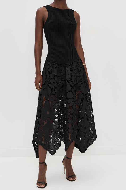 A woman wearing a sleeveless midi dress featuring a fitted ribbed bodice and a sheer, intricately patterned floral lace skirt with an asymmetrical hem.