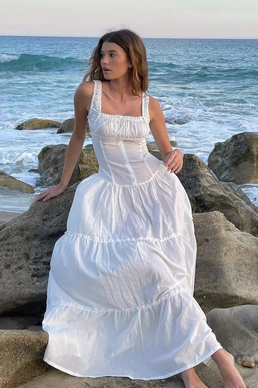 White - A woman sitting on rocks by the ocean wearing a white, sleeveless maxi dress featuring a ruched square neckline, corset-style buttoned bodice, and a voluminous tiered skirt.