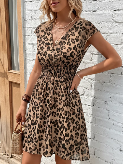 Leopard - Woman modelling a lightweight animal-print v-neck mini dress with cap sleeves and a shirred waist, holding a woven bag against a white brick wall.