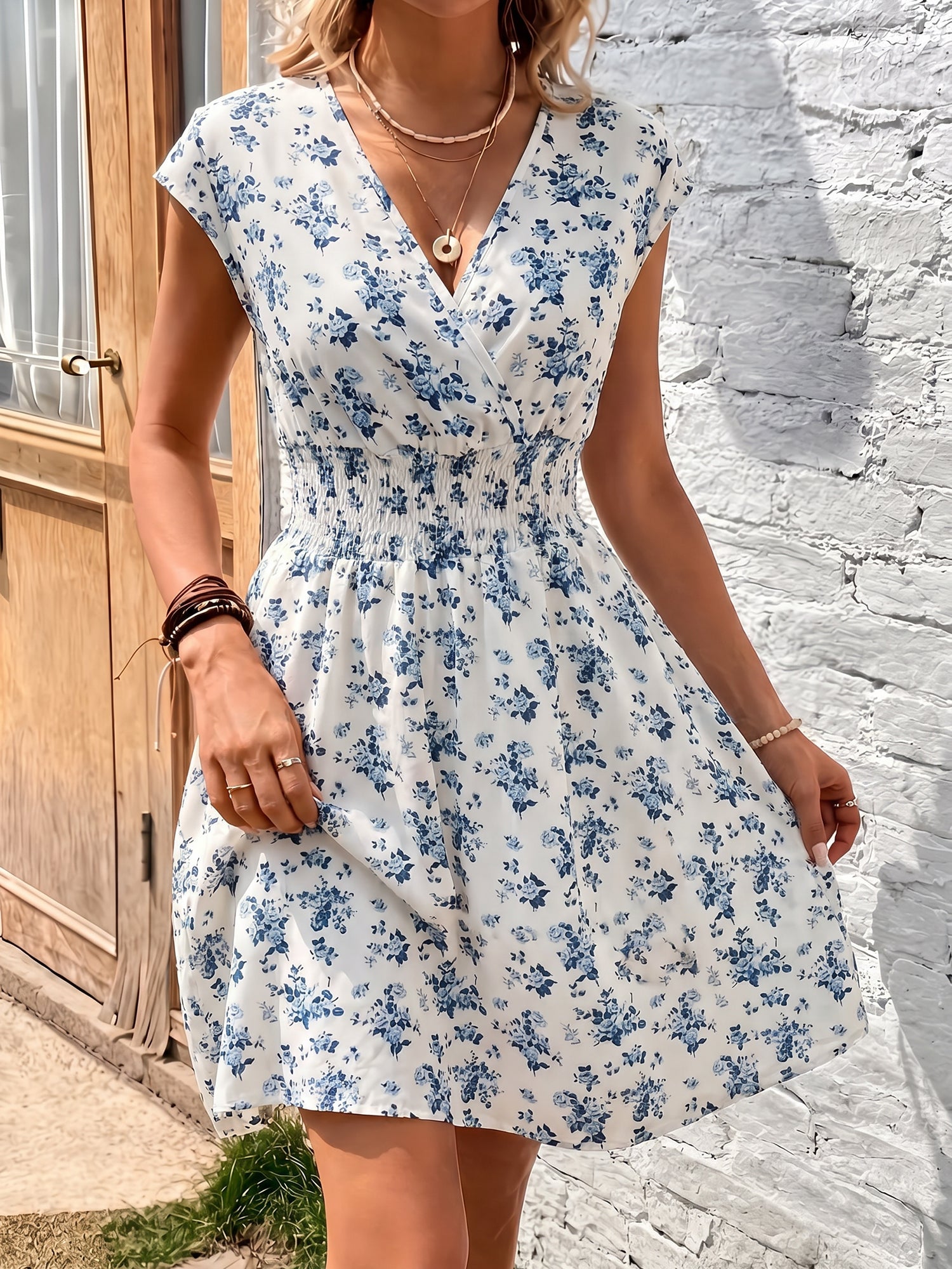 Blue - Woman modelling a lightweight animal-print v-neck mini dress with cap sleeves and a shirred waist, holding a woven bag against a white brick wall.