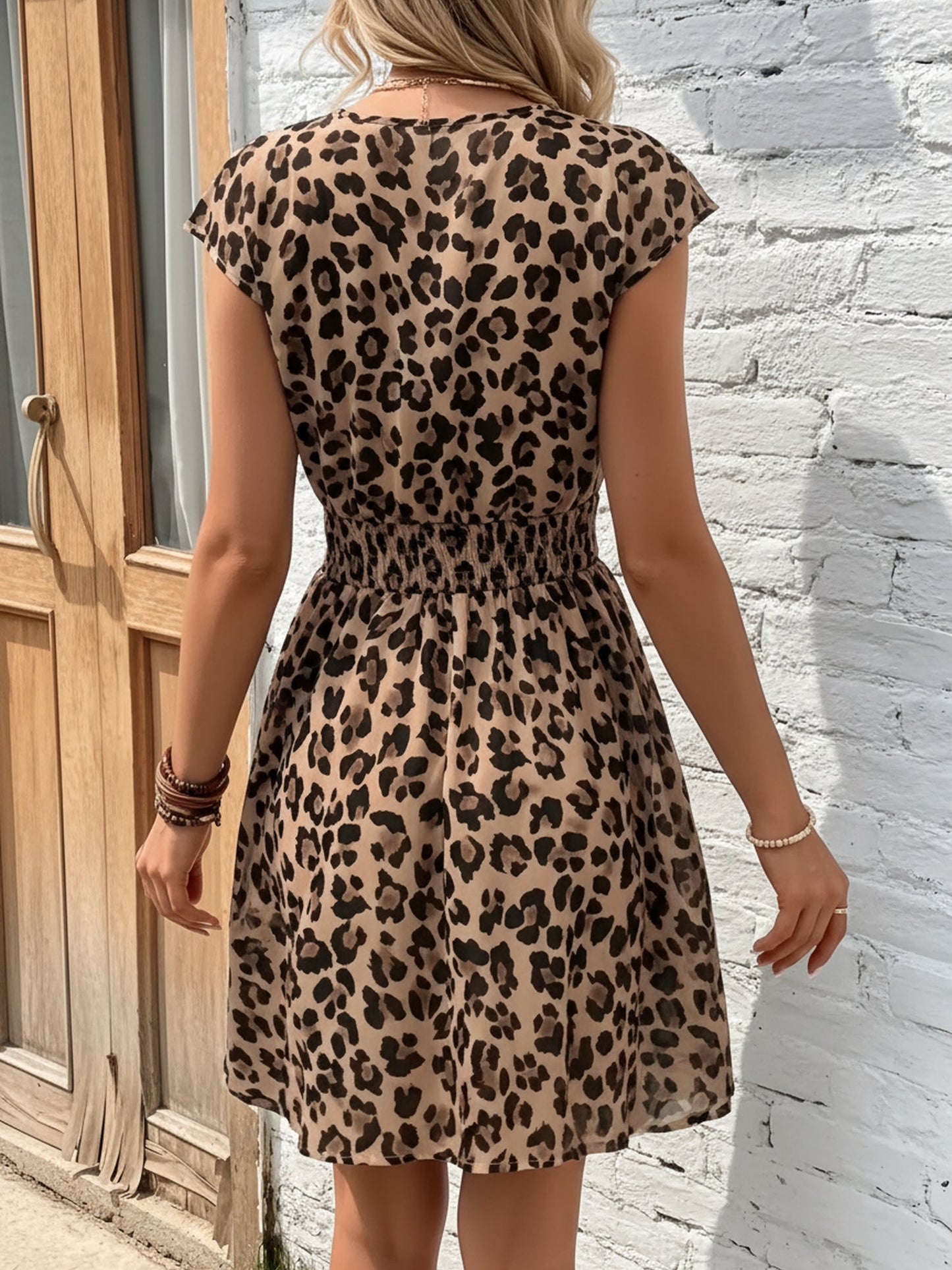 Leopard - Woman modelling a lightweight animal-print v-neck mini dress with cap sleeves and a shirred waist, holding a woven bag against a white brick wall.