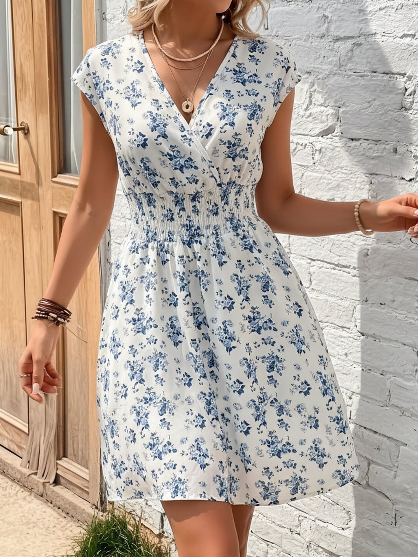 Blue - Woman modelling a lightweight animal-print v-neck mini dress with cap sleeves and a shirred waist, holding a woven bag against a white brick wall.