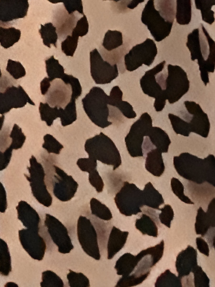 Leopard - Woman modelling a lightweight animal-print v-neck mini dress with cap sleeves and a shirred waist, holding a woven bag against a white brick wall.