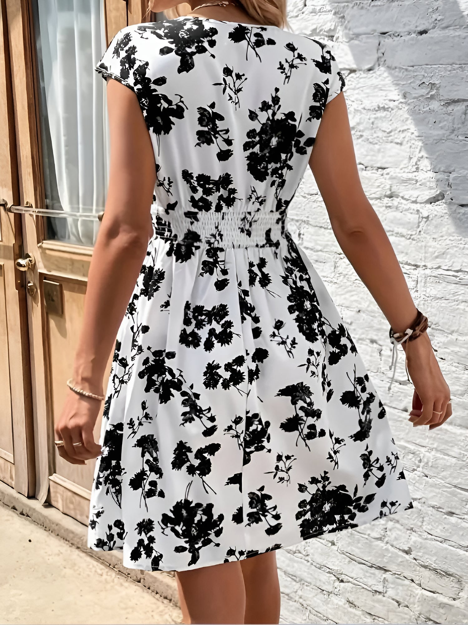 White - Woman modelling a lightweight animal-print v-neck mini dress with cap sleeves and a shirred waist, holding a woven bag against a white brick wall.