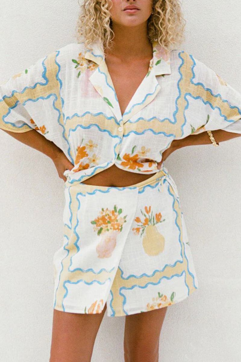 A woman modelling a matching white cropped short-sleeve shirt and mini wrap skirt, decorated with a yellow and blue wavy border pattern and orange floral vase illustrations.