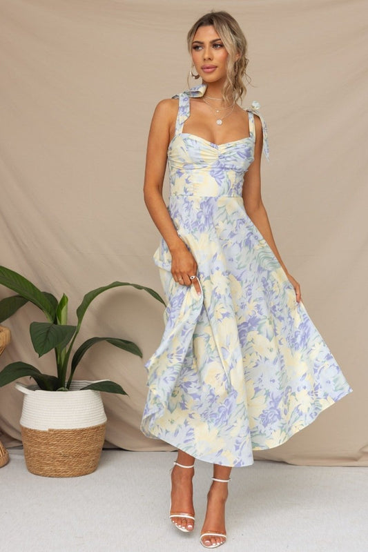 Blue - A woman models a pastel floral midi dress with a ruched sweetheart bodice and tie-shoulder straps. The A-line skirt is light blue, yellow, and lavender. She is wearing white heels and standing against a beige background.