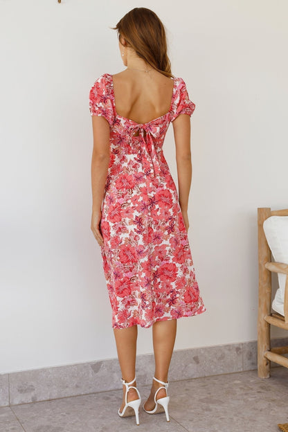 A woman wearing a vibrant pink and red floral midi dress with a sweetheart neckline, short puff sleeves, and a high side slit.