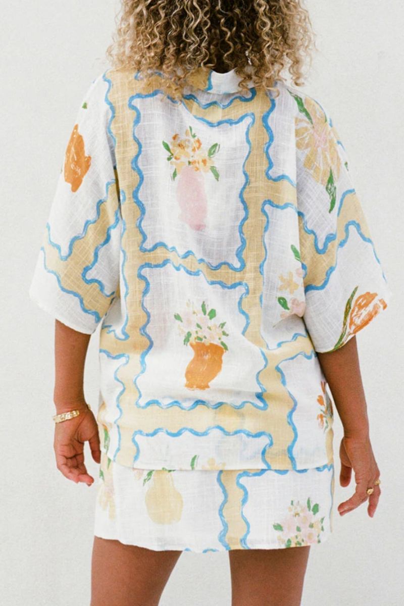 MultiColor - A woman modelling a matching white cropped short-sleeve shirt and mini wrap skirt, decorated with a yellow and blue wavy border pattern and orange floral vase illustrations.