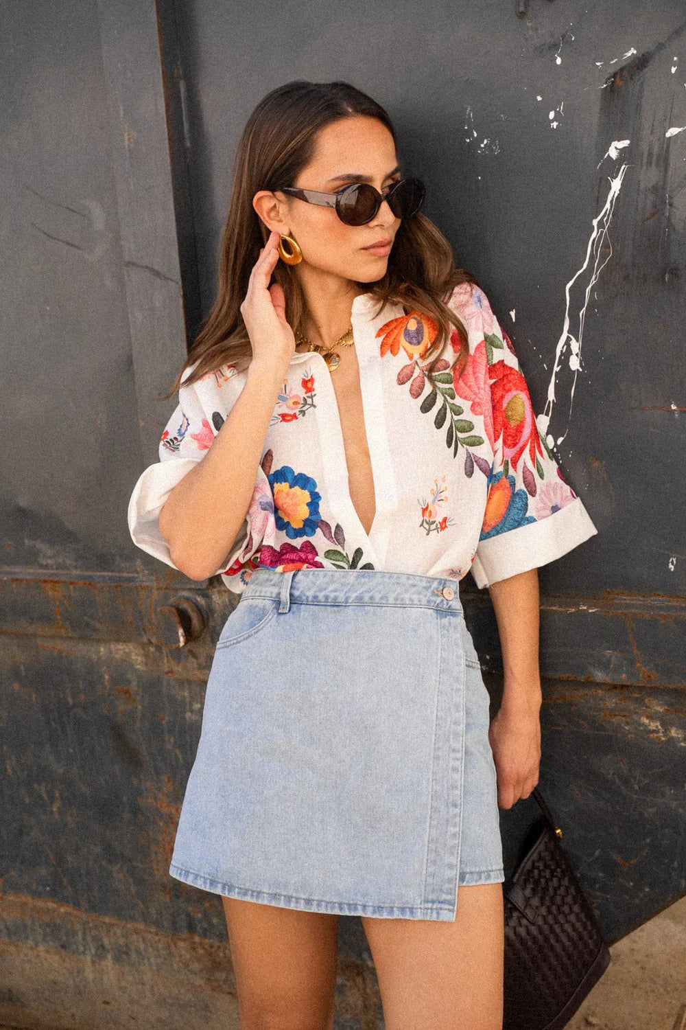 A woman wearing a white, relaxed-fit button-down blouse featuring vibrant, large-scale multicoloured floral embroidery, styled with rolled sleeves and tucked into a khaki bottom.