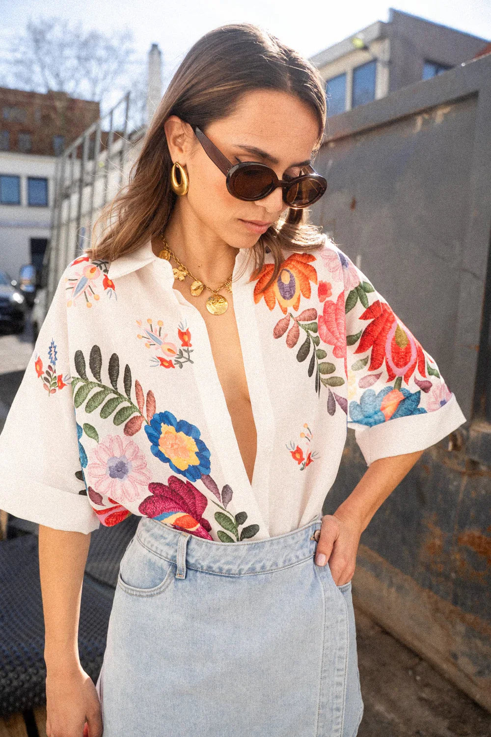 Print - A woman wearing a white, relaxed-fit button-down blouse featuring vibrant, large-scale multicoloured floral embroidery, styled with rolled sleeves and tucked into a khaki bottom.
