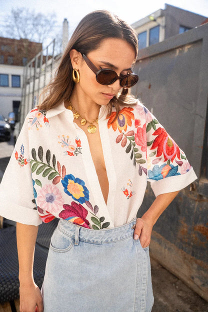 Print - A woman wearing a white, relaxed-fit button-down blouse featuring vibrant, large-scale multicoloured floral embroidery, styled with rolled sleeves and tucked into a khaki bottom.