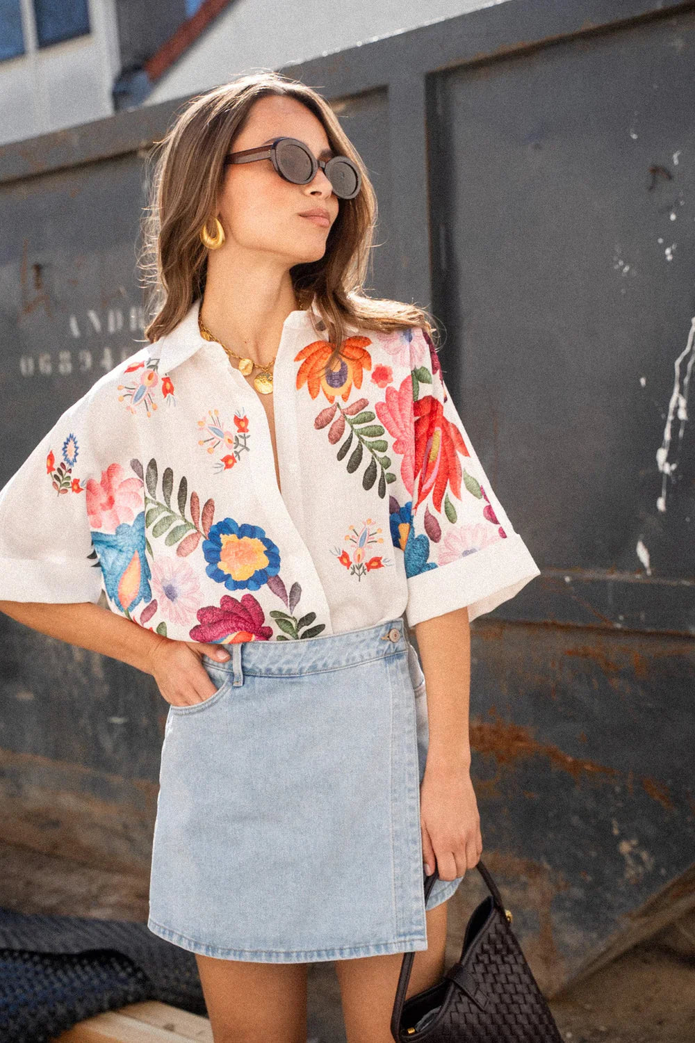 Print - A woman wearing a white, relaxed-fit button-down blouse featuring vibrant, large-scale multicoloured floral embroidery, styled with rolled sleeves and tucked into a khaki bottom.
