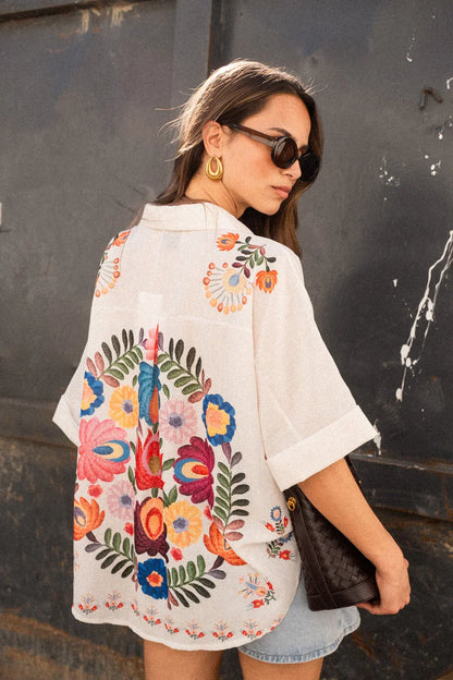 Print - A woman wearing a white, relaxed-fit button-down blouse featuring vibrant, large-scale multicoloured floral embroidery, styled with rolled sleeves and tucked into a khaki bottom.