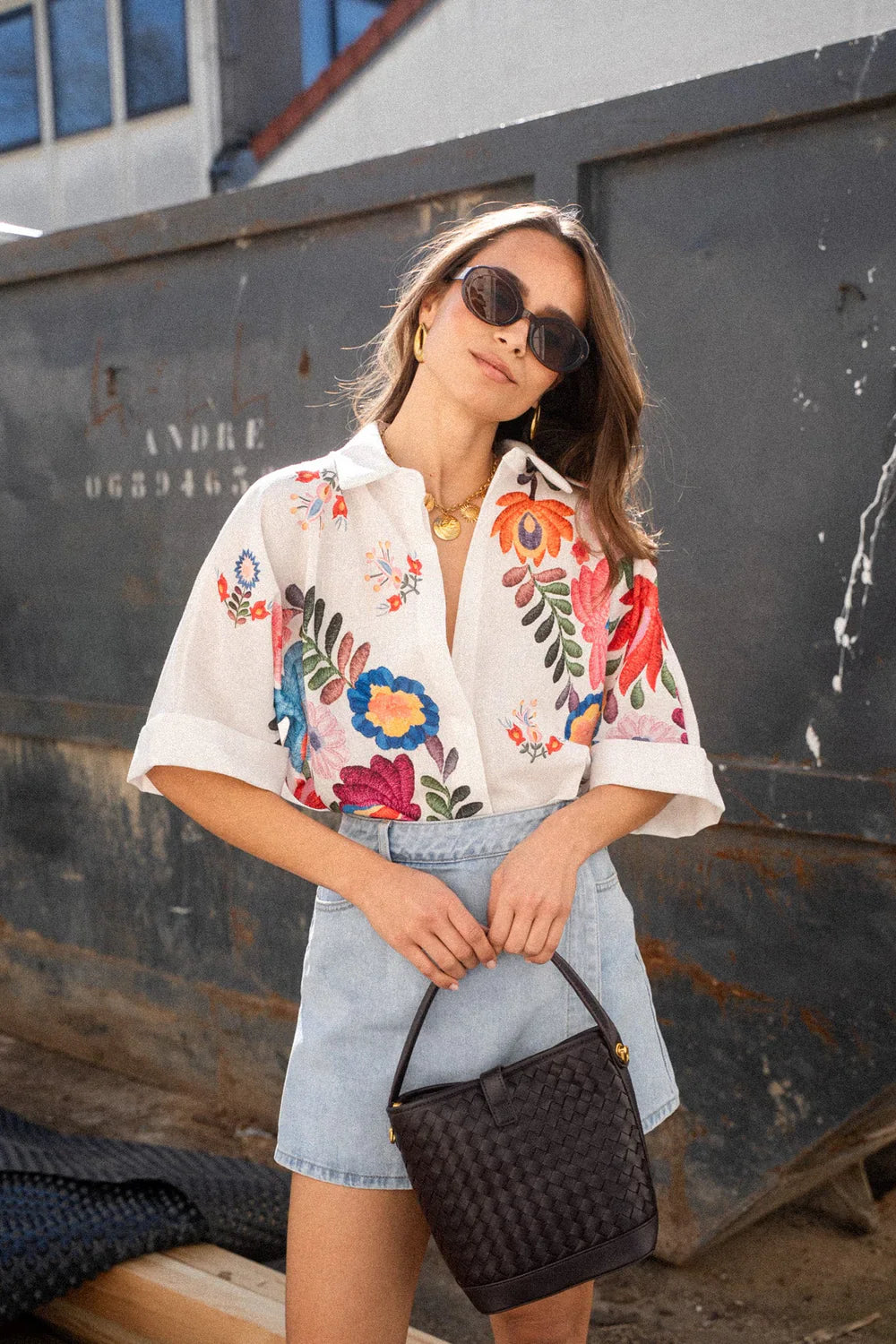 Print - A woman wearing a white, relaxed-fit button-down blouse featuring vibrant, large-scale multicoloured floral embroidery, styled with rolled sleeves and tucked into a khaki bottom.