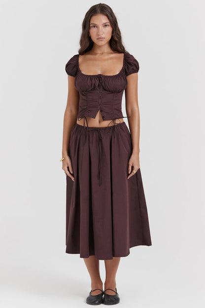 A woman models a dark brown midi dress featuring a ruched, button-front bodice with cap sleeves, daring waist cutouts with tie details, and a gathered flowing skirt, standing outdoors by rustic wooden doors.
