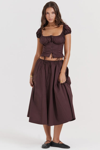 Brown - A woman models a dark brown midi dress featuring a ruched, button-front bodice with cap sleeves, daring waist cutouts with tie details, and a gathered flowing skirt, standing outdoors by rustic wooden doors.