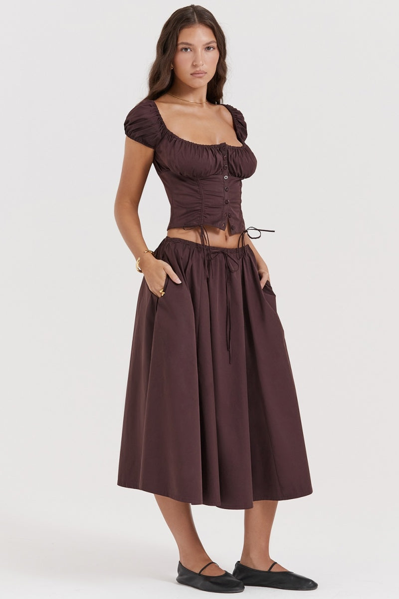 Brown - A woman models a dark brown midi dress featuring a ruched, button-front bodice with cap sleeves, daring waist cutouts with tie details, and a gathered flowing skirt, standing outdoors by rustic wooden doors.