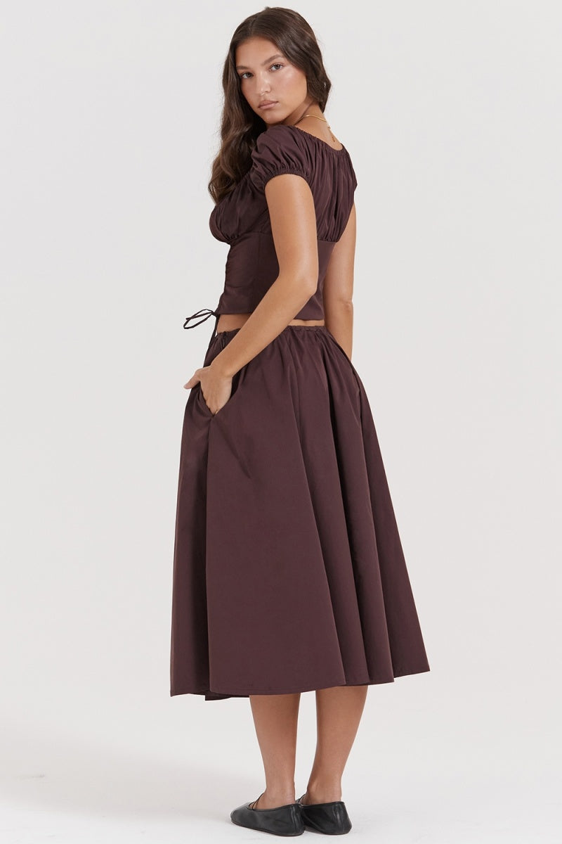 Brown - A woman models a dark brown midi dress featuring a ruched, button-front bodice with cap sleeves, daring waist cutouts with tie details, and a gathered flowing skirt, standing outdoors by rustic wooden doors.