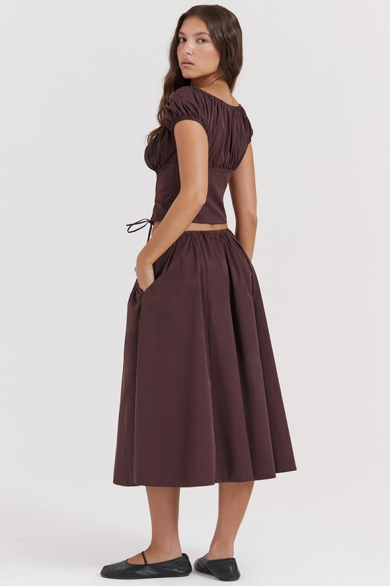 Brown - A woman models a dark brown midi dress featuring a ruched, button-front bodice with cap sleeves, daring waist cutouts with tie details, and a gathered flowing skirt, standing outdoors by rustic wooden doors.