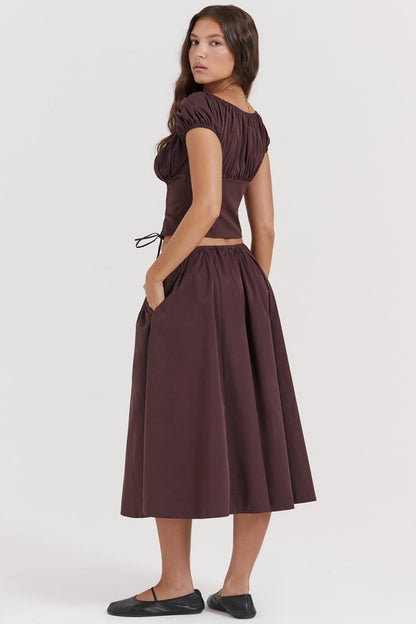 Brown - A woman models a dark brown midi dress featuring a ruched, button-front bodice with cap sleeves, daring waist cutouts with tie details, and a gathered flowing skirt, standing outdoors by rustic wooden doors.