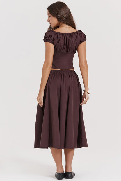 Brown - A woman models a dark brown midi dress featuring a ruched, button-front bodice with cap sleeves, daring waist cutouts with tie details, and a gathered flowing skirt, standing outdoors by rustic wooden doors.