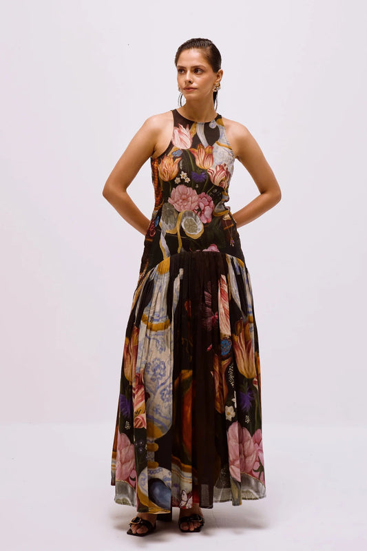 Multi - Full-length view of a woman wearing a sleeveless maxi dress with a high halter neckline and a pleated skirt, featuring a detailed still life floral and vase print on a dark background.