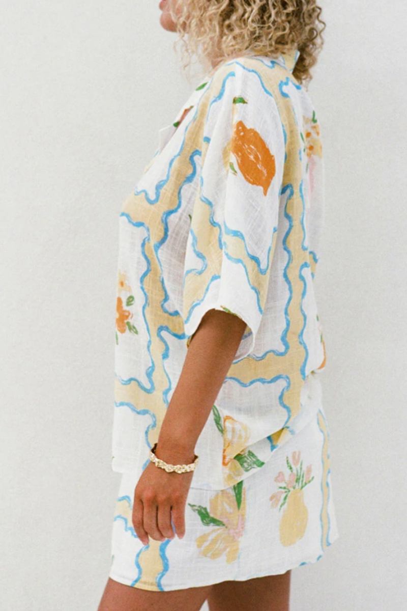 MultiColor - A woman modelling a matching white cropped short-sleeve shirt and mini wrap skirt, decorated with a yellow and blue wavy border pattern and orange floral vase illustrations.