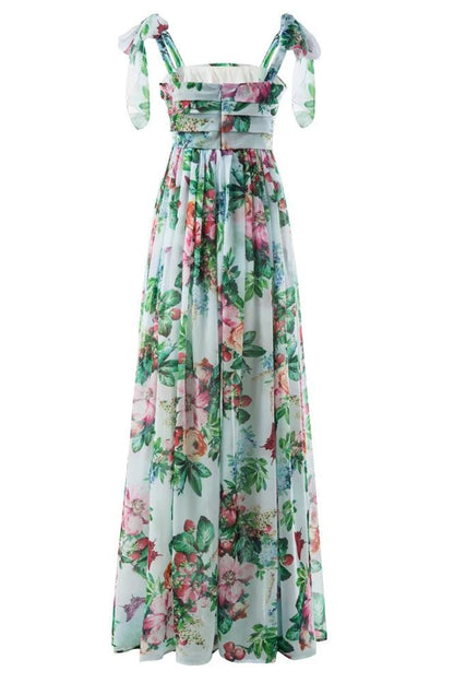 Multi - A woman models a flowing maxi dress featuring a vibrant, large-scale botanical floral print, styled with a ruched bodice and wide tie-shoulder straps, standing outdoors near a floral arrangement.